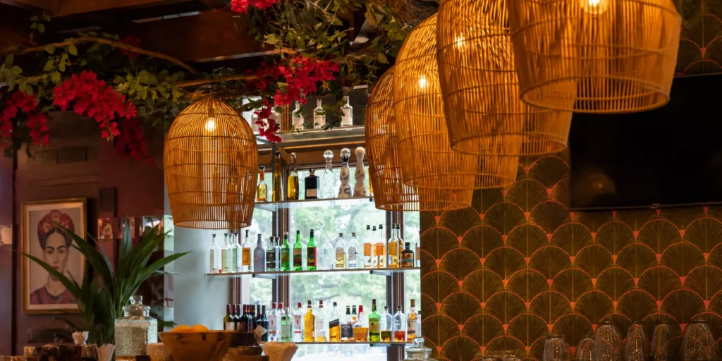 View of a bar with four rows of bottle, hanging lamps and a portrait of Mexican painter Fridda.